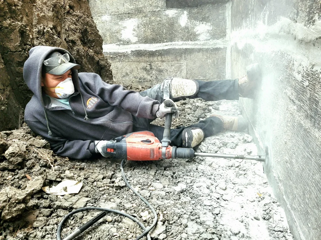 Excavator carving a trench alongside a residential foundation for drainage repair