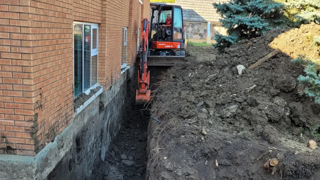 Excavator carving a trench alongside a residential foundation for drainage repair