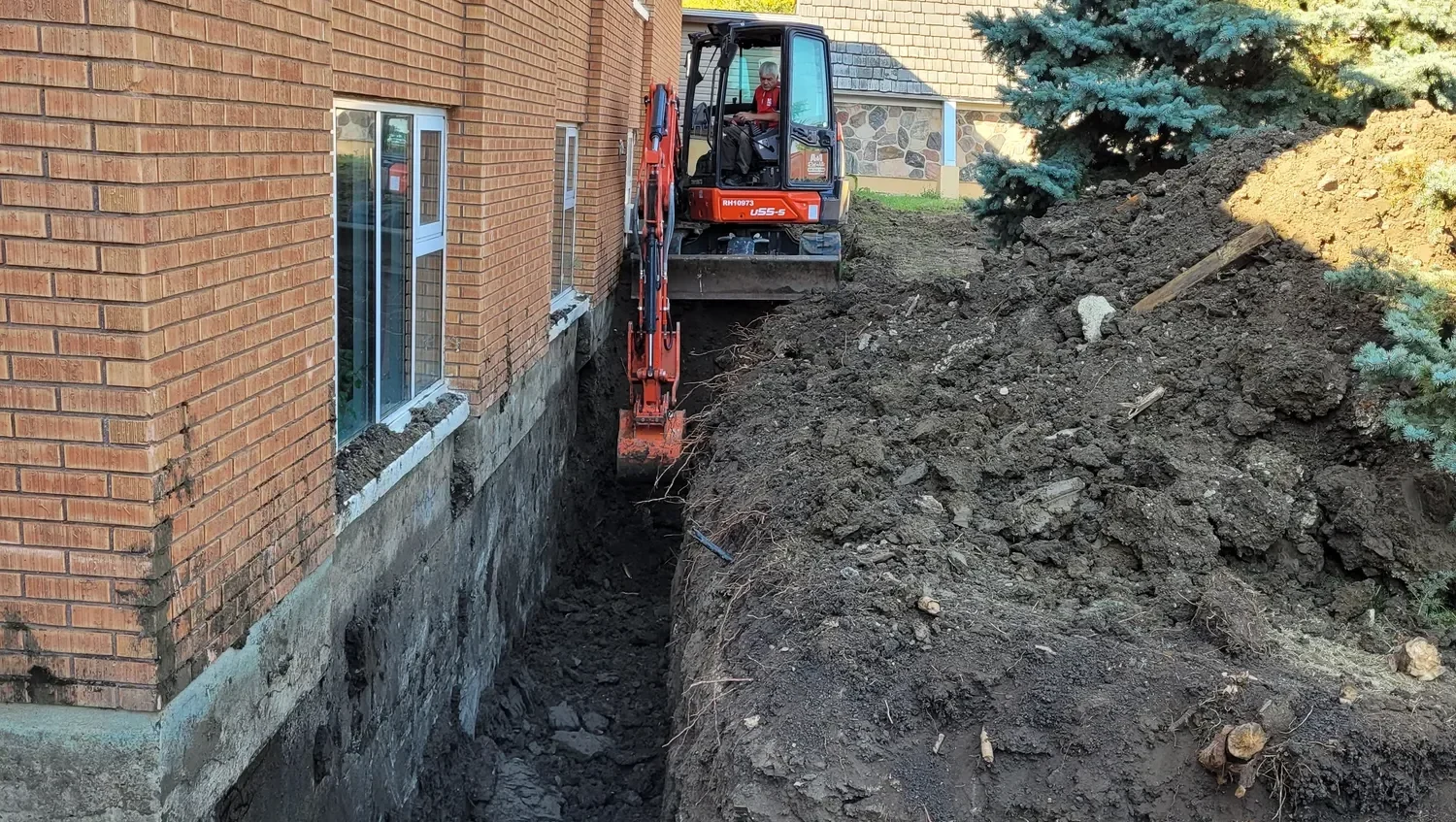 Excavator carving a trench alongside a residential foundation for drainage repair